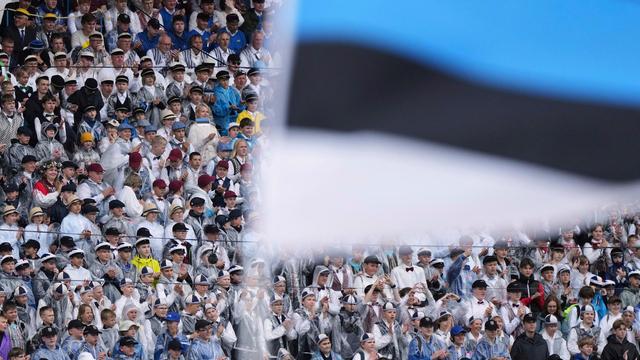 Chorsänger sitzen beim jährlichen Liederfestival in Tallinn in einem Stadion. Vor ihnen im Bildvordergrund wird eine estnische Flagge geschwenkt.