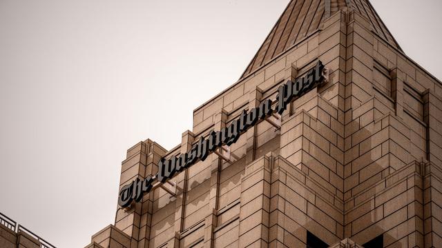 Close-up of The Washington Post logo on a beige stone building with a pointed roof against a clear, light sky.
