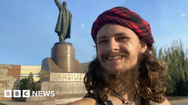 A man with long hair and a beard stands in front of a Soviet statue