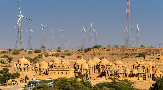 Sandsteinbauten in der Wüste von Jaisalmer vor einer Reihe moderner Windkraftanlagen.