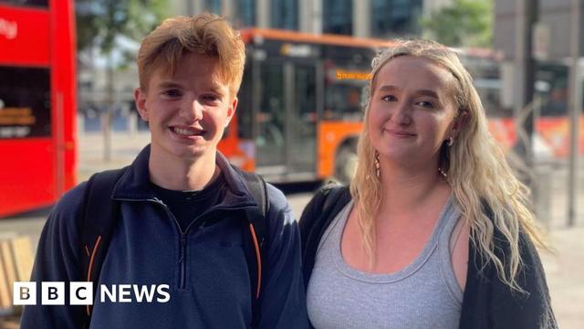 Students Evan Stoddart and Holly Wells, who stand in front of Wood Street near Central Square, Cardiff. Evan (left) has ginger short hair, and wears a dark blue quarter zip fleece and a backpack, while Holly (right) has blonde long hair and wears a grey vest and cardigan. There are buses in the background, which are blurred as the two people, in the foreground are the focus of the picture.