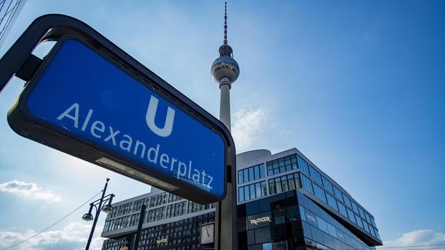 Aufnahme des U-Bahn Schildes am Bahnhof Alexanderplatz. Im Hintergrund ist vor blauem Himmel auch der Fernsehturm zu sehen.