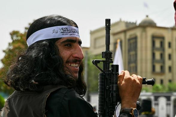 Nahaufnahme: Ein Taliban mit langem schwarzen Haar das von einer weißen Binde mit islamistischen Slogan beschrieben ist, dreht sich über die Schulter, lächelend, sein Gewehr auf Gesichtshöhe in den Himmel zeigend