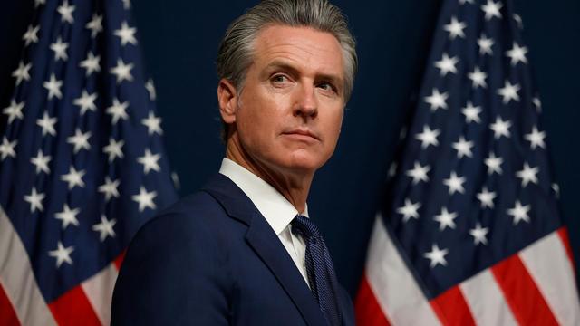 Kaliforniens Gouverneur Gavin Newsom trägt einen blauen Anzug und steht zwischen zwei amerikanischen Flaggen.