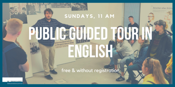 Ein Museumsguide erklärt in einer Ausstellung Dinge. Hinter ihm eine Wand mit Fotos. Eine Gruppe sitzende und stehende Menschen hören ihm zu. 