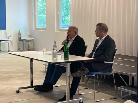Ein Tisch mit zwei Männern nebeneinander. Der eine Herr links ist älter mit weißem Haar. Der rechts jünger, es ist der Dolmetscher. 