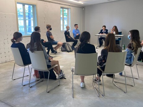 Eine Gruppe junger Menschen auf Stühlen im Kreis, am Ende sitzt eine ältere Dame an einem Tisch, neben ihr eine jüngere Frau mit langem dunklen Haar, sie ist die Dolmetscherin. 