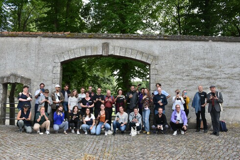 Vor einer Mauer mit einem Tor stehen ind hocken Menschen zu einem Gruppenfoto. Sie halten alle ihre Handys in der Hand. 