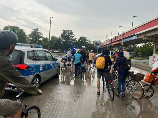 Mehrere Menschen auf dem Rad stehen am Berliner Eastgate und warten auf den Beginn der Demo. Links ein Polizeiauto zu sehen.