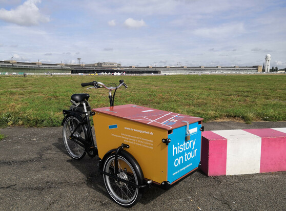 Ein Foto eines bunten Lastenrades auf dem Tempelhofer Feld. Im HIntergrund sieht man das ehemalige Flughafengebäude. Das Lastenrad hat vorn eine hellblaue Fläche, auf der "history on Tour" in großen Buchstaben steht. Oben kann man einen Grundriss auf der Fläche erkennen. Die der Kamera zugewandte Seite ist ockerfarben und enhält neben der Website ns-zwangsarbeit.de die Adresse und Öffnungszeiten des Dokumentationszentrums NS-Zwangsarbeit. 