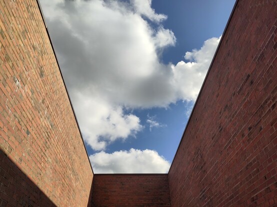 In den blauen Himmel blickend, mit weiß-Grauen Wolken. Der Blick ist eingeschränkt durch drei hohe Ziegelmauern, die die Gefangenschaft im Lager symbolisieren.