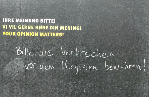 Tafel mit der Aufforderung "Ihre Meinung bitte!", mit Kreide beantwortet: "Bitte die Verbrechen vor dem Vergessen bewahren!"