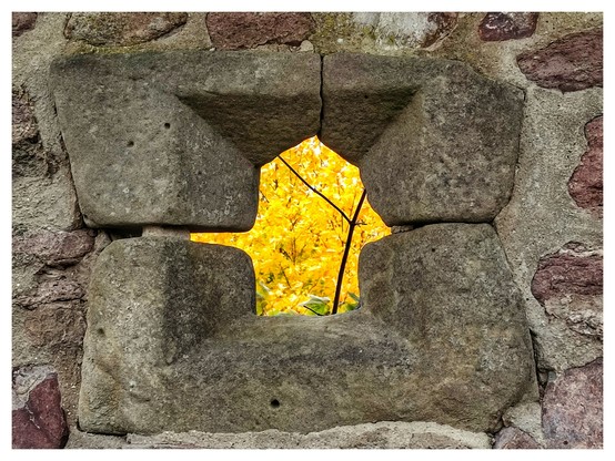Blick durch ein kleines Fenster in einer alten Schlossmauer. Das Fenster ist aus großen Steinen gebaut und hat eine unregelmäßige Form. Im Hintergrund steht ein Baum mit gelben Herbstblättern, die durch das Fenster zu sehen sind, so dass der Durchblick komplett gelb aussieht. 