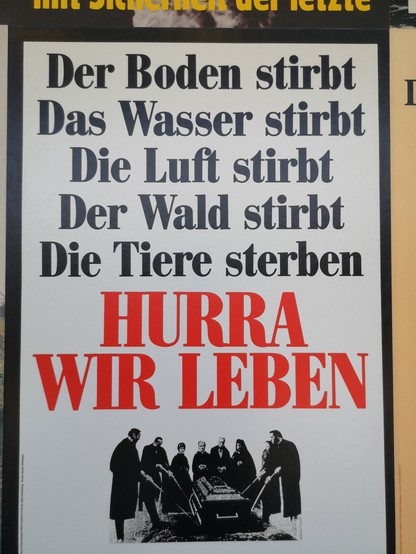 Ein Plakat von Klaus Staeck mit der Aufschrift:
Der Boden stirbt 
Das Wasser stirbt 
Die Luft stirbt 
Der Wald stirbt 
Die Tiere sterben 
HURRA WIR LEBEN

Darunter eine Grabszene, wo Personen einen Sarg in die Erde hinablassen. 