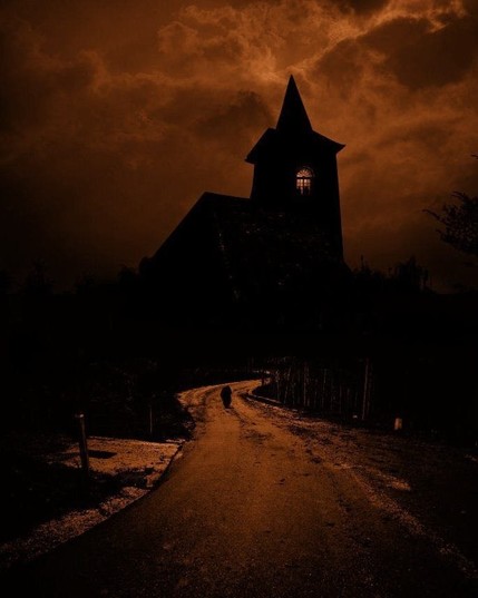 A photo in black and dark red.

A road with a lone person walking on a road owards a small church.

There is a lit window in the tower, where two human silhouettes can be seen.
