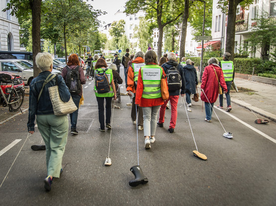 Fußverkehrsaktive spazieren auf der Straße und ziehen jeweils einen Schuh hinter sich her.
