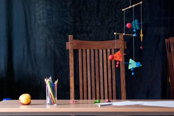 Buntfoto: Holzstuhl hinter Tisch mit Bunt- und Wachsmalstiften, einer nekatrine und papier. Darüber hängt ein buntes Mobile . Foto und Copyright: John Colton