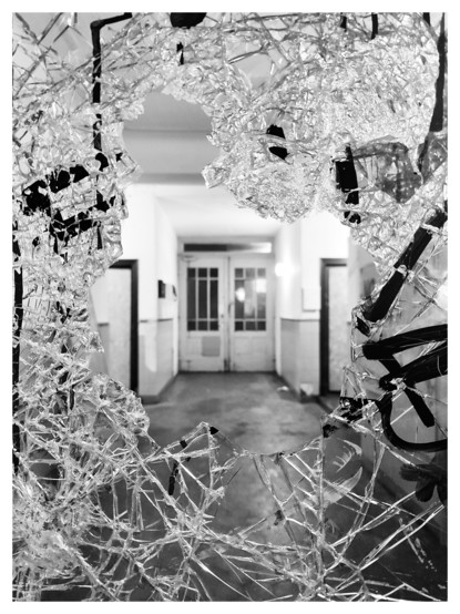 Schwarz-Weiß-Foto. Wir schauen durch die zerbrochene Glasscheibe einer Hauseingangstür in einen Hausflur. Die Reste der Scheibe bilden ein scharfes und gezacktes Loch. Im Hausflur ist Licht an, am Ende des Flures sieht man die Tür zum Hof, sie ist unscharf. 
