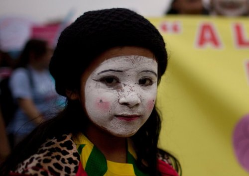 A young woman at a march.

It's a portrait. She's looking at the camera. Her face is painted white, with cracks, some rouge on her lips and cheeks.

She looks sad and yet determined.

She's wearing a black hat and colourful clothes. Behind her, blurred, other women marching.
