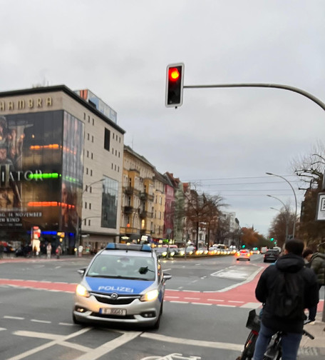 Das Bild zeigt eine städtische Kreuzung bei bewölktem Wetter. Im Vordergrund fährt ein Polizeiauto mit eingeschaltetem Licht in Richtung Kamera, auf einem Radweg steht ein Fahrradfahrer mit dunkler Kleidung. Links im Hintergrund ist ein großes Gebäude mit einer leuchtenden Reklame für ein Kino (Alhambra) zu sehen. Die Ampel oben zeigt rotes Licht. Entlang der Straße stehen Autos und bunte Altbauten, während sich weiter hinten die Straße in die Ferne erstreckt.