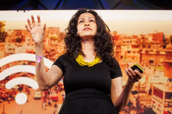 Payal Arora steht auf der Stage 1 vor einem großen Bildschirm, auf dem ein Foto einer Stadt zu erkennen ist, außerdem ein WIFI-Symbol. Sie trägt ein dunkles Kleid und eine gelbe Halskette.