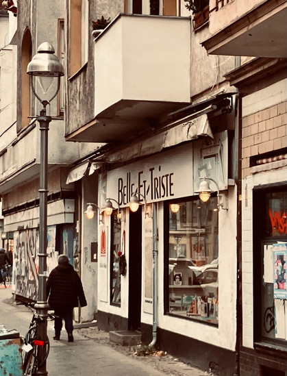 Ein Laden in einer Berliner Straße mit der Aufschrift "Belle et Triste" auf dem Schild. Vor dem Schaufenster hängt ein Kranz, und im Inneren des Ladens sind Bücher und andere Gegenstände zu sehen. Die Fassade wirkt etwas gealtert, daneben sind Graffiti auf den Wänden. Eine Person geht an der Szenerie vorbei.