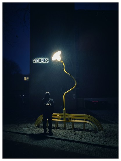 Nächtliche Szene mit kühlem Blaustich. Hinter einer interessant gebogenen Straßenlaterne mit gelbem Mast, die an einer gelben Bank befestigt ist, steht ein Mann. Er ist nur von hinten und in Umrissen zu sehen. Es schneit leicht, das kann man im Lichtkegel der Laterne sehen. Hinter der Laterne und dem Mann ist an einem dunklen Haus ein leuchtendes Schild zu sehen, darauf steht St. Agnes. 