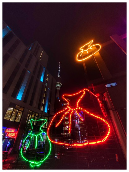Nachtaufnahme. Drei bunte Neonschilder bzw. Umrisse aus gebogenen Neonleuchtstäben in Form von Teigtaschen scheinen in der Luft zu hängen. Im Hintergrund ist der Fernsehturm zu erahnen. 