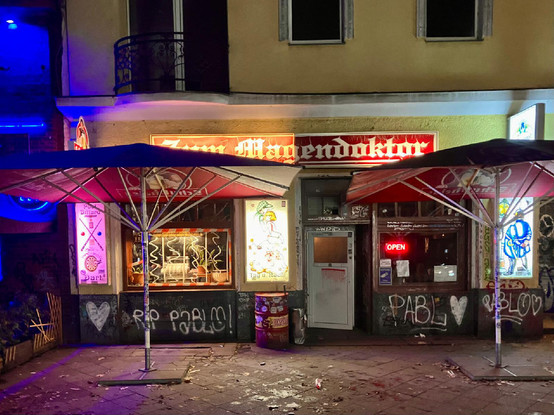 Außenansicht der Kneipe "Zum Magendoktor" in Berlin-Wedding bei Nacht. Der Schriftzug leuchtet in roter Neonbeleuchtung, daneben ein "Open"-Schild. Vor dem Eingang stehen zwei rote Sonnenschirme, die Fassade ist mit Graffiti bedeckt. Auf dem Boden liegen Blätter und Müll.

