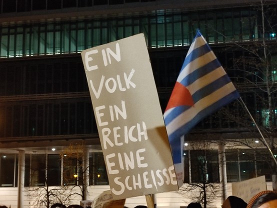 Bild eines Demoschildes, darauf steht "Ein Volk, ein Reich, eine Scheiße". Dahinter eine Flagge des VVN-BDA mit blauen und weißen Streifen und einem roten, auf der Spitze stehenden Dreieck. 