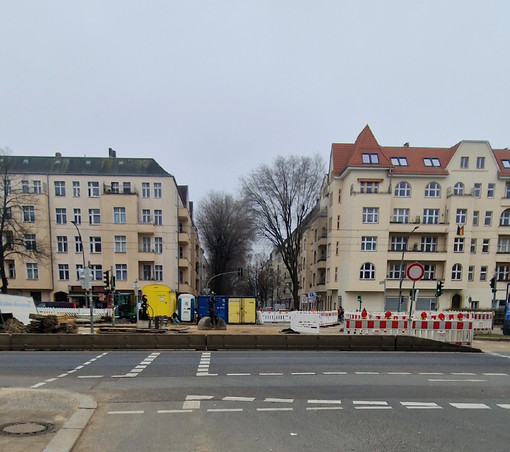 Das Bild zeigt eine Straßenszene mit einer Baustelle in einem städtischen Gebiet, umgeben von typischen Berliner Altbauten.

