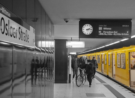 U-Bahnhof Osloer Straße in Berlin-Wedding: Gelber U-Bahnzug der Linie U8 steht am Bahnsteig. Passagiere steigen ein und aus, einige gehen am Bahnsteig entlang. Ein Schild mit dem Stationsnamen und eine analoge Uhr sind sichtbar.

