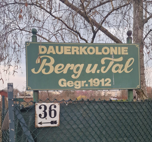 Grünes Schild mit der Aufschrift „Dauerkolonie Berg u. Tal Gegr. 1912“ an einem Zaun; darunter ein kleines Schild mit der Hausnummer 36 und einem Pfeil nach links. Im Hintergrund Bäume und Wohnhäuser.

