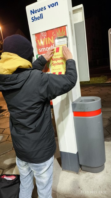 Ein Mensch steht nachts vor einer Ankündigungstafel von Shell und beklebt sie mit einem Flyer: "Kaputtes System"