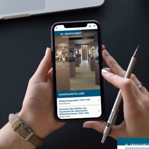 Das Bild zeigt eine Person, die ein Smartphone hält. Auf dem Display ist der Multimediaguides des Dokumentationszentrums NS-Zwangsarbeit zu sehen. Es werden zwei Dauerausstellungen angezeigt: „Alltag Zwangsarbeit 1938-1945“ (Dauer 60 min) und „Zwischen allen Stühlen. Die Geschichte der italienischen Militärinternierten 1943-1945“ (Dauer 30 min).