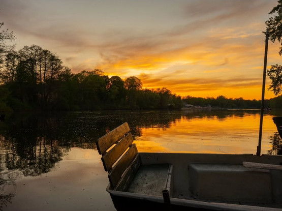 Ein Ruderboot mit Holzsitz am Ufer des Plötzensees in Berlin-Wedding bei Sonnenuntergang, mit Spiegelungen der Bäume und des orangefarbenen Himmels auf der Wasseroberfläche.

