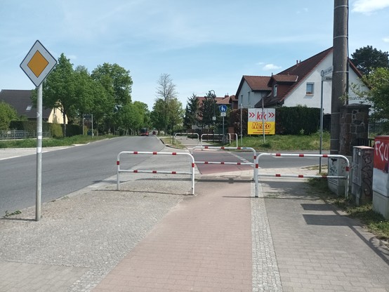 Geh- und Radweg in ländlicher Gegend, Wohngebiet, direkt von einer Einmündung von rechts. mit drei rotweißen Drängelgittern wird das geradeaus laufen und auch radfahren unmöglich gemacht. Fußys müssen auf den Radweg treten und umgekehrt. GeradeausfahrendeAutos haben Vorfahrt, abbiegende dadurch auch.
