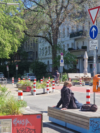 Straßenszene in einem umgestalteten Stadtbereich mit Sitzbänken, Pflanzkästen, Blumen und Verkehrspollern; Menschen nutzen die Sitzgelegenheiten bei sonnigem Wetter, im Hintergrund Wohnhäuser und Bäume.