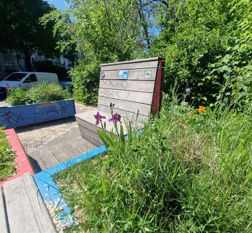 Straßenszene in einem umgestalteten Stadtbereich mit Sitzbänken, Pflanzkästen, Blumen und Verkehrspollern; Menschen nutzen die Sitzgelegenheiten bei sonnigem Wetter, im Hintergrund Wohnhäuser und Bäume.