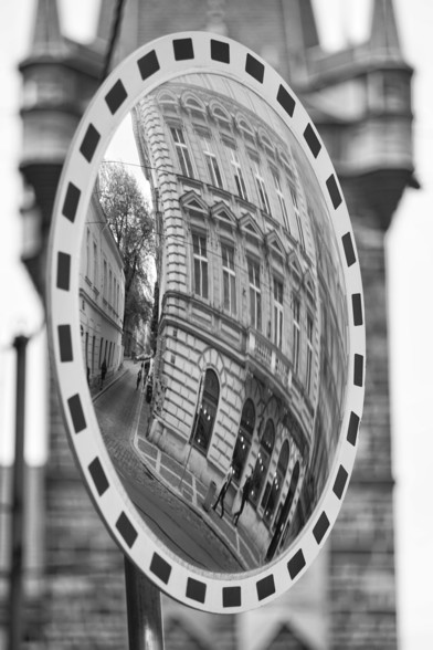 Schwarz-Weiß-Foto im Hochformat. Im Hintergrund unscharf ein Turm, im Vordergrund ein Verkehrsspiegel, in dem sich ein Prager Altbau spiegelt.