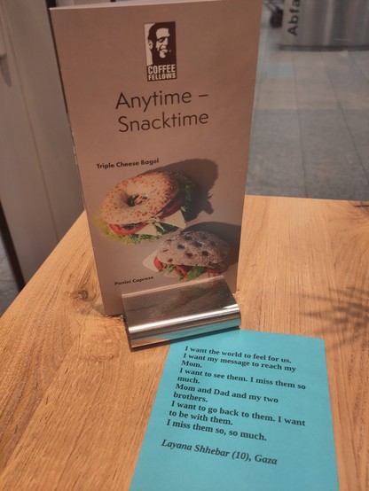 A Coffeefellows menu with the headline "anytime - snacktime" standing on a table. In front of it a blue paper with the following text.

I want the world to feel for us.
I want my message to reach my Mom.
I want to see them. I miss them so much.
Mom and Dad and my two brothers.
I want to go back to them. I want to be with them.
I miss them so, so much.

Layana Shhebar (10), Gaza
