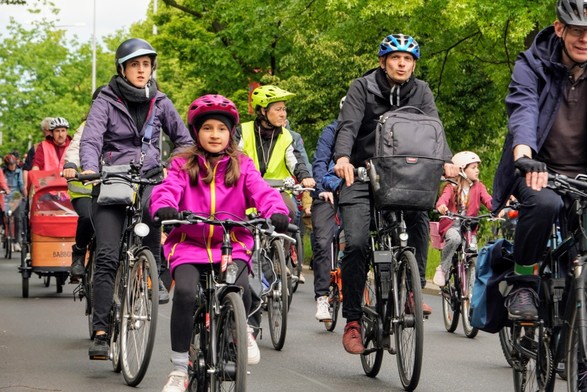 Viele Menschen fahren auf einer großen Straße Fahrrad. Sie haben bunte Sachen an und sind unterschiedlich alt: vom Kindesalter zu ihren Eltern bis hin zu Senioren.