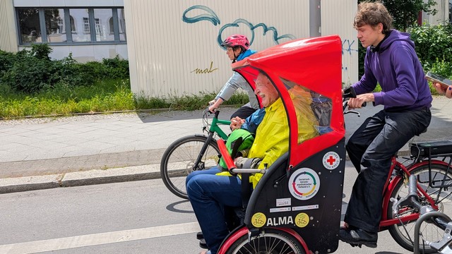 Das Bild zeigt eine Szene auf einer Straße, in der ein Mann in einem roten und schwarzen Fahrradkarren mit einem älteren lachenden Herrn. Der ältere Herr trägt einen gelben Regenmantel und sitzt im Karren, während das Kind in einem grünen Helm und einem blauen Regenmantel sitzt. Der Mann am Lenker des Fahrrads trägt ein lila Sweatshirt und schaut nach vorne. Neben ihnen fährt ein weiterer Radfahrer mit einem grünen Fahrrad. Im Hintergrund ist ein Gebäude mit Graffiti zu sehen, und die Straße ist asphaltiert. Die Szene wirkt entspannt .