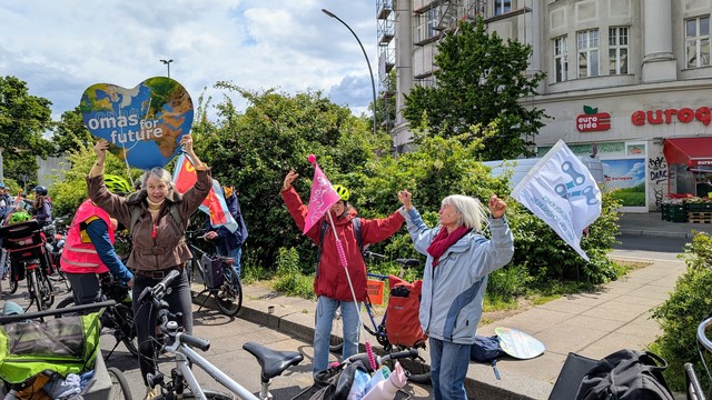 Das Bild zeigt eine Gruppe von Menschen, die an einer Demonstration teilnehmen. Im Vordergrund stehen zwei Frauen, die fröhlich mit erhobenen Armen und Fahnen tanzen. Eine Frau hält ein Herzförmiges Schild mit der Aufschrift "Omas for future" hoch, während die andere eine rosa Fahne mit einem Symbol hält. Beide Frauen tragen Jacken und sind umgeben von Fahrrädern. Im Hintergrund sind weitere Demonstranten zu sehen, einige mit Fahrrädern und Kinderwagen. Die Szene spielt an einem sonnigen Tag in einer städtischen Umgebung.
