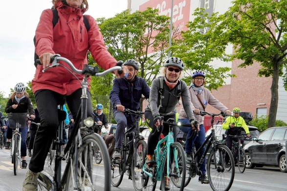 Das Bild zeigt eine Gruppe von Radfahrern, die auf einer Straße in einer städtischen Umgebung fahren. Vorderste Radfahrerin trägt eine rote Jacke und einen Helm und fährt eine klassische Fahrradart mit einem Lenker. Neben ihr fährt ein Mann in einem dunkelblauen Jackett und Helm, gefolgt von einer Frau in einem grauen Sweatshirt und Helm, die lächelt. Weitere Radfahrer sind im Hintergrund zu sehen, einige tragen Helme und leuchtendfarbene Kleidung. Die Straße ist asphaltiert und von Bäumen gesäumt, die Blätter zeigen, dass es Frühling oder Sommer ist. Im Hintergrund ist ein Gebäude mit einem großen Schild zu sehen, das teilweise den Text "Häuser" enthält. Ein Polizist in einem gelben Sicherheitsweste und Helm fährt ebenfalls mit, um die Radfahrer zu begleiten. 