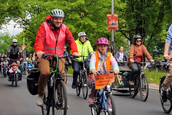 Das Bild zeigt eine Gruppe von Menschen, die auf Fahrrädern durch eine grüne Straße fahren. Im Vordergrund ist ein Mann in einem roten Anorak und einem Helmlächelnd auf seinem Fahrrad. Neben ihm fährt ein Kind in einem pinken Helm und einem orangefarbenen Westen auf einem blauen Fahrrad, das ein Schild mit der Aufschrift "ICH WILL ALLEINE ZUR SCHULE FAHREN KÖNNEN" trägt. Im Hintergrund sind weitere Fahrradfahrer zu sehen, darunter Kinder in Kinderwagen und Erwachsene in verschiedenen Outfits. 