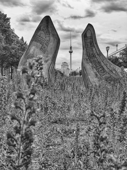 Schwarz-Weiß-Foto im Hochformat. Zu sehen ist ein Kunstwerk aus zwei bearbeiteten Steinen, die auf dem Mittelstreifen der Frankfurter Allee zwischen sehr vielen Lavendelpflanzen stehen. Das Bild ist aus einer sehr tiefen Perspektive aufgenommen, wenn man durch die Steine hindurchschaut, sieht man den Fernsehturm und eins der Hochhäuser am Strausberger Platz. 