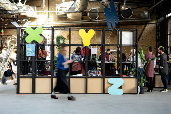 Auf dem Foto ist eine Trennwand aus Quadern zu sehen. Dahinter befindet sich der Makerspace auf der re:publica mit einigen Teilnehmenden. Im Vordergrund läuft eine Person vorbei. Über und vor ihr befinden sich die bunten Buchstaben X, Y und Z.