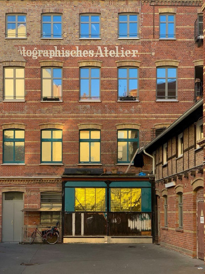 Backsteinfassade eines mehrstöckigen Altbaus mit der verblassten Aufschrift „Photographisches Atelier“, darunter ein Hof mit einem gelbverglasten Anbau, davor ein abgestelltes Fahrrad.

