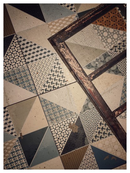 Hochformatiges Farbfoto. Es zeigt verschiedene Fliesen auf einem Badezimmerboden. Die haben bräunliche, grünliche und beige Muster, sind aber etwas alt und abgeschabt. 